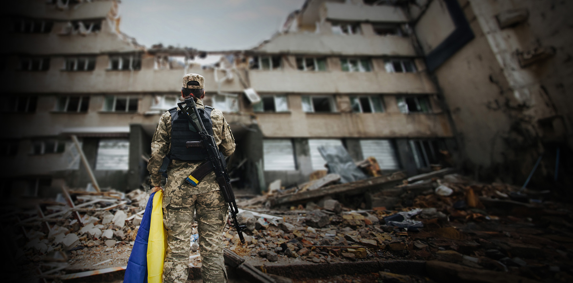 Kvinnlig soldat som håller en Ukraina flagga, med ryggen vänd mot kameran och tittar på en förstörd byggnad i Ukraina.